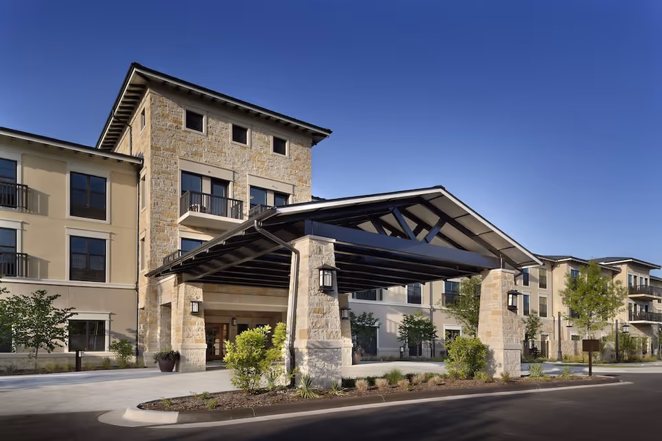 Exterior view of Belmont Village Senior Living West Lake Hills building with a covered entrance supported by stone pillars, surrounded by landscaped greenery and clear blue sky.