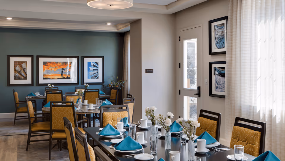 A well-lit dining room with multiple tables set for a meal, featuring yellow cushioned chairs, turquoise napkins, white plates, cups, and glassware. The room has a teal accent wall adorned with framed artwork and a door with windows letting in natural light through sheer curtains.