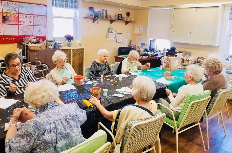 A group of elderly women sitting around a large table in a well-lit room, engaging in a craft activity painting flower pots. The room has windows with blinds, shelves with decorations, and various supplies on the table.