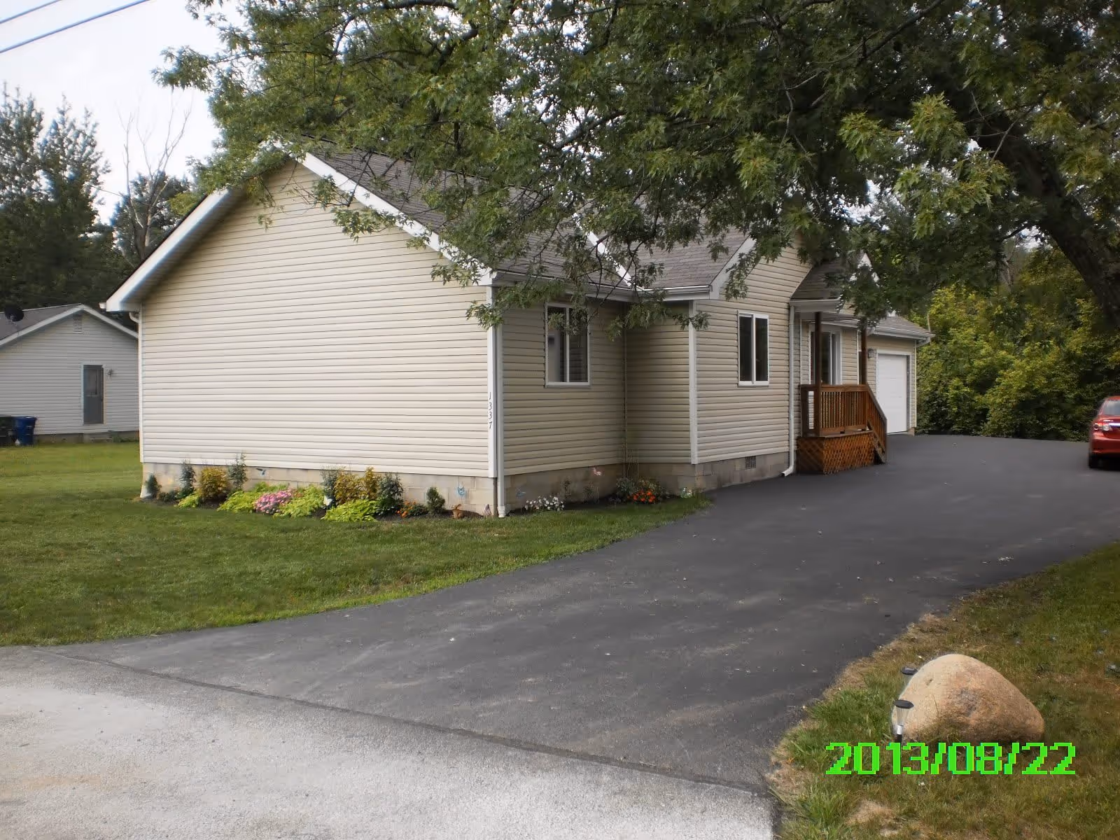Exterior view of a single-story beige house with a sloped roof, a small wooden porch, and a driveway leading to a garage. The house is surrounded by green grass, some bushes, and trees. A red car is parked at the end of the driveway. The date 2013/08/22 is displayed in green text at the bottom right corner.