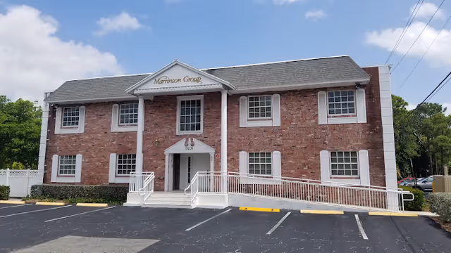 Two-story brick building with white trim and a gray shingled roof. The entrance has white columns and a ramp leading up to the door. The building has multiple windows and is surrounded by a parking lot with marked spaces. Trees and a partly cloudy sky are visible in the background.