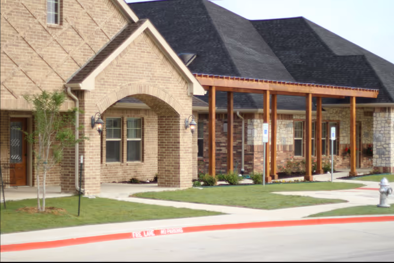Exterior view of a senior living facility building with brick and stone walls, a covered walkway supported by wooden beams, green lawn, and a fire hydrant near the curb marked as a fire lane with no parking.
