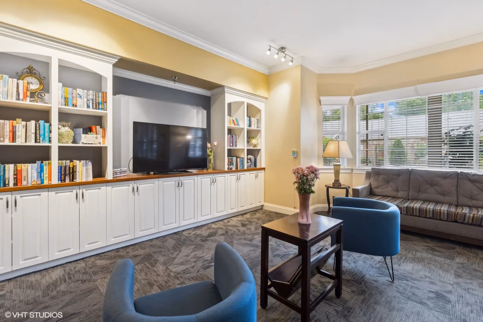 A cozy living room with a large built-in white bookshelf and cabinets, a flat-screen TV, two blue armchairs, a wooden side table with a pink vase of flowers, a gray couch, a side table with a lamp, and large windows with white blinds letting in natural light.