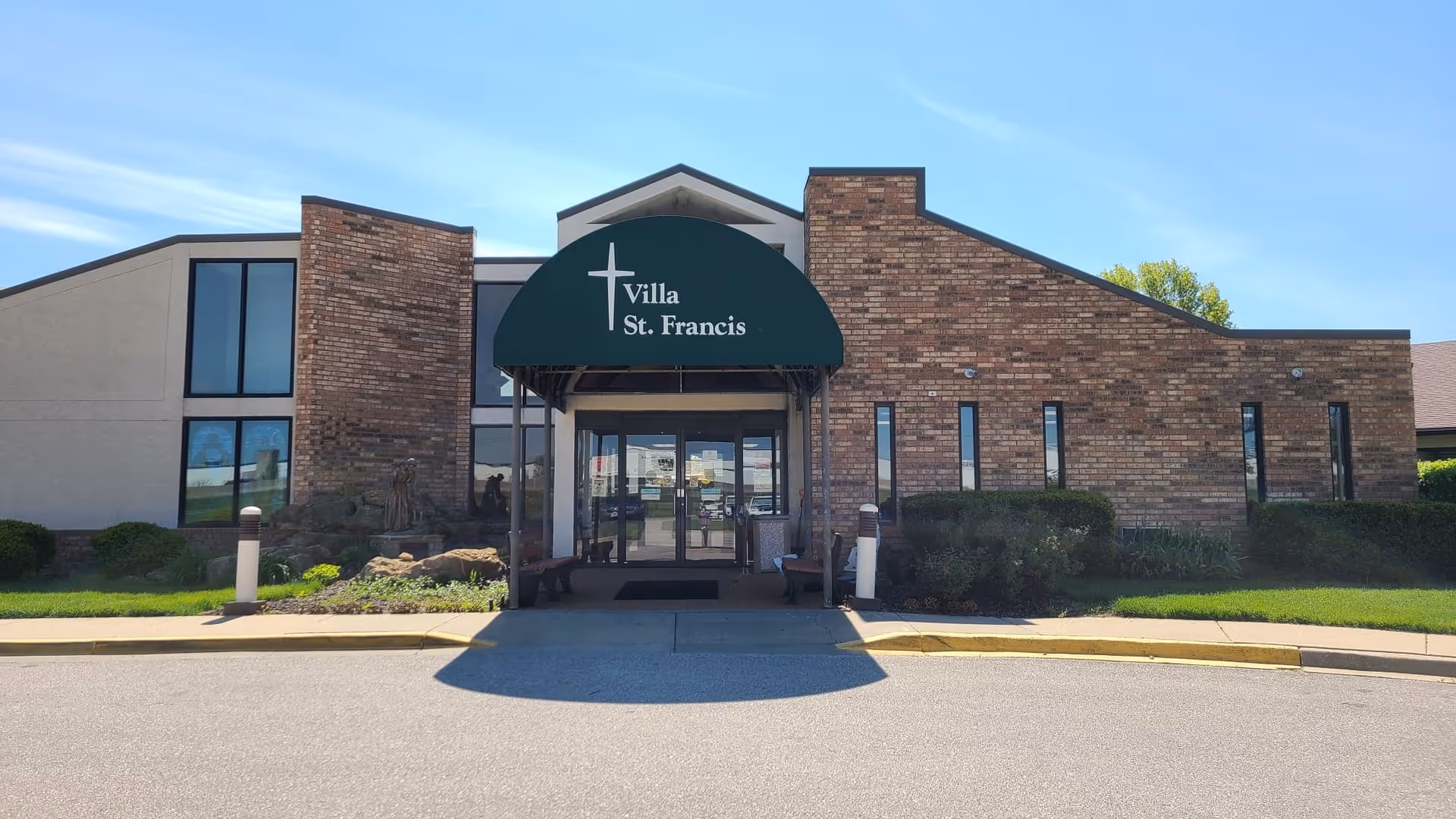 Front exterior view of Villa St. Francis Catholic Care Center building with a brick facade, large windows, and a green awning displaying the facility name and a cross symbol above the entrance.