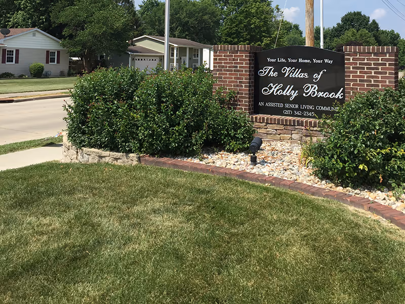 A brick sign for The Villas of Holly Brook assisted senior living community surrounded by green bushes and landscaping rocks, with a grassy lawn and residential houses in the background under a clear sky.