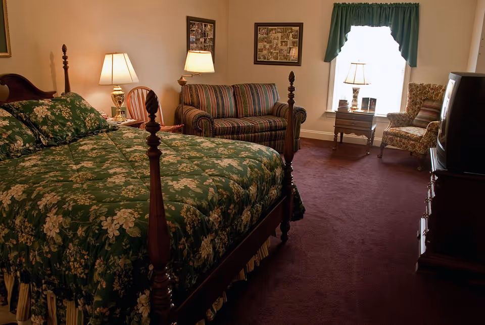 A cozy bedroom featuring a four-poster bed with green floral bedding, a striped sofa and armchair, lamps, a TV, and a window with a valance.