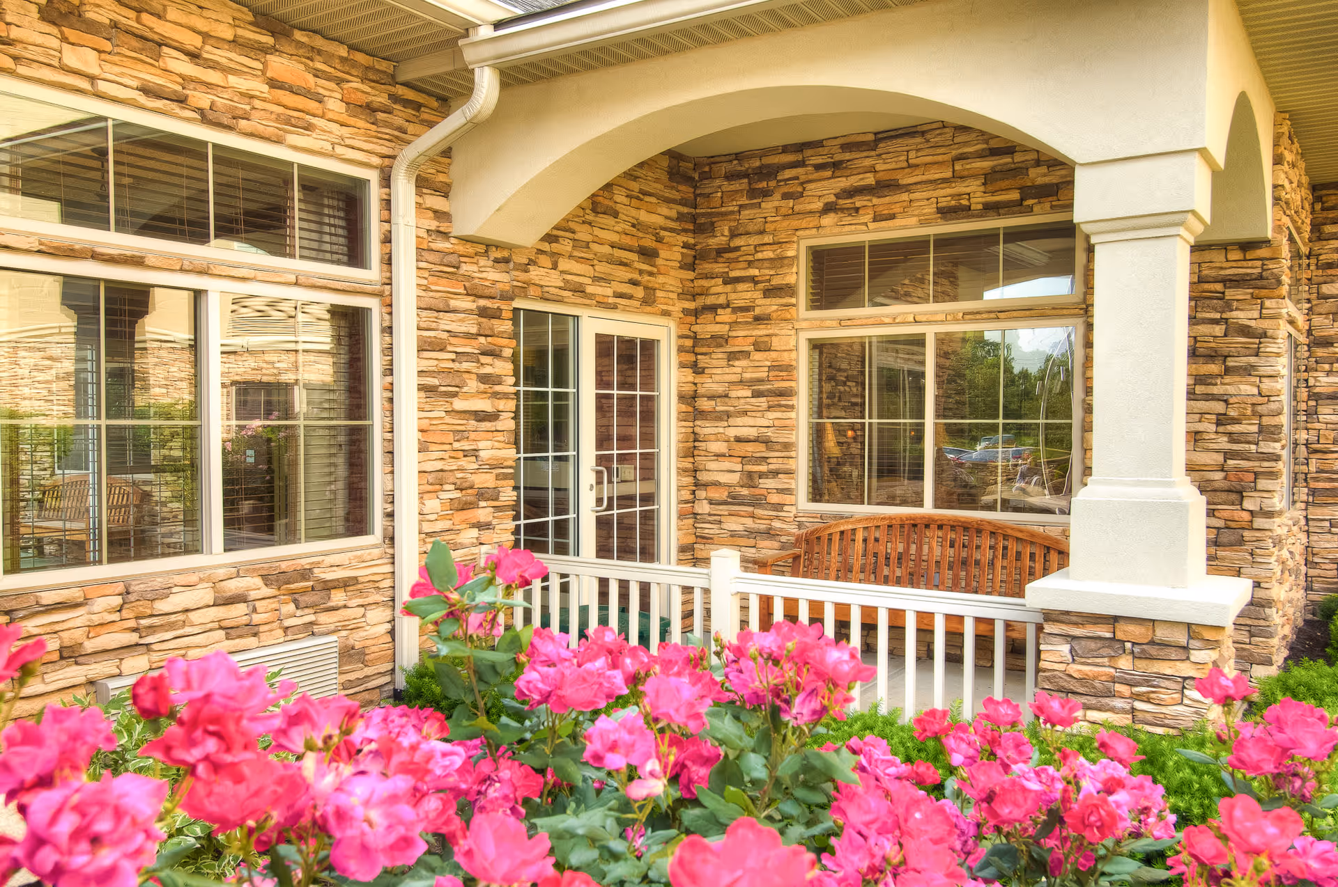 Outdoor patio area of Westside Garden Plaza with stone walls, large windows, a wooden bench, white railing, and vibrant pink flowers in the foreground.