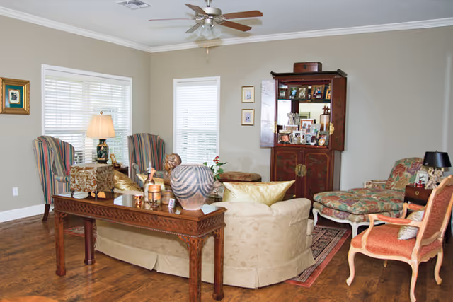 A warm living room with a curved beige sofa, patterned armchairs, a wooden console table, decorative cabinet and ceiling fan over hardwood floors.