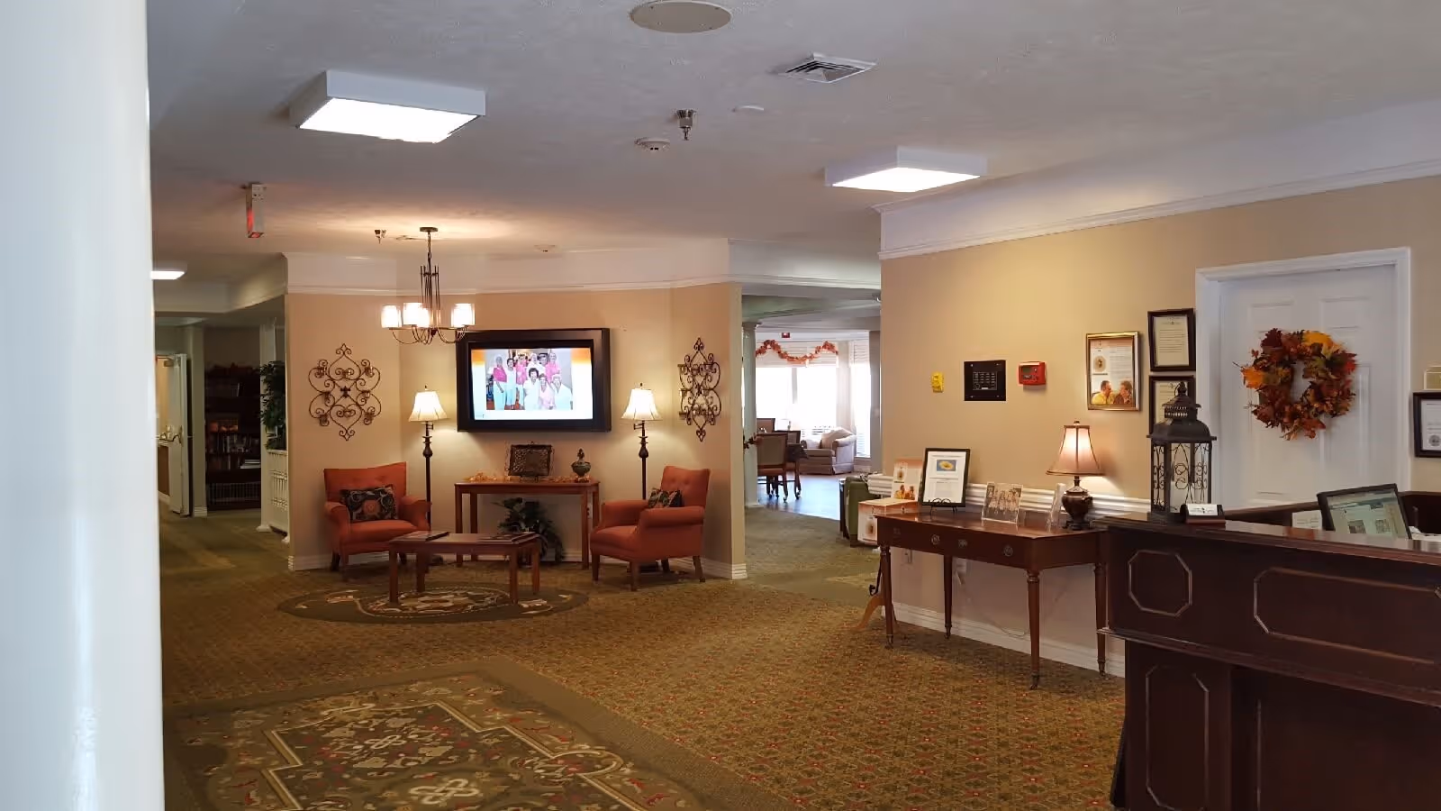 Interior view of a senior living facility lobby area with patterned carpet, two orange armchairs, a wooden coffee table, a wall-mounted TV displaying a group photo, two standing lamps, a chandelier, and a reception desk with a computer. The walls are decorated with framed pictures and a fall-themed wreath on a door.
