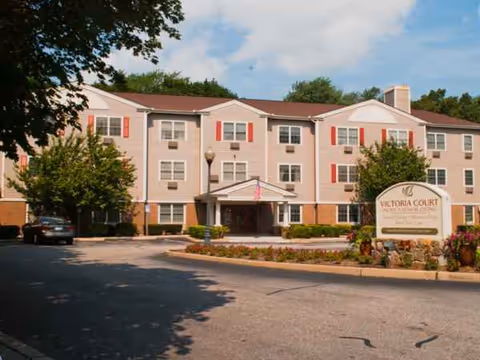 Exterior view of Victoria Court Memory Care, a beige multi-story building with red shutters.