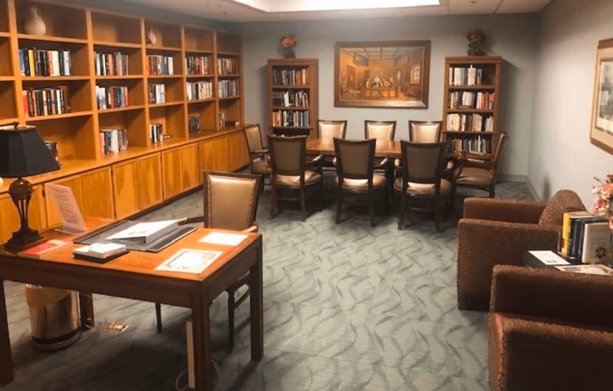 A cozy library or reading room with wooden bookshelves filled with books along the left wall and two smaller bookshelves at the back. There is a large wooden table with eight chairs in the center of the room, a wooden desk with a chair and lamp on the left side, and two brown upholstered armchairs on the right. The room has carpeted flooring and framed artwork on the walls.