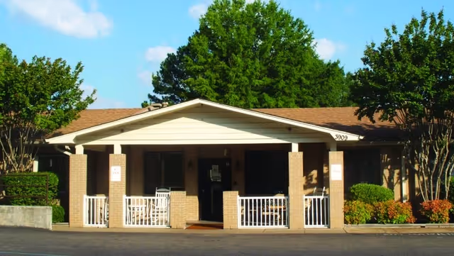 Front exterior view of a single-story building with a covered porch supported by brick columns. The porch has white railings and rocking chairs. The building is surrounded by green trees and shrubs under a blue sky with some clouds.
