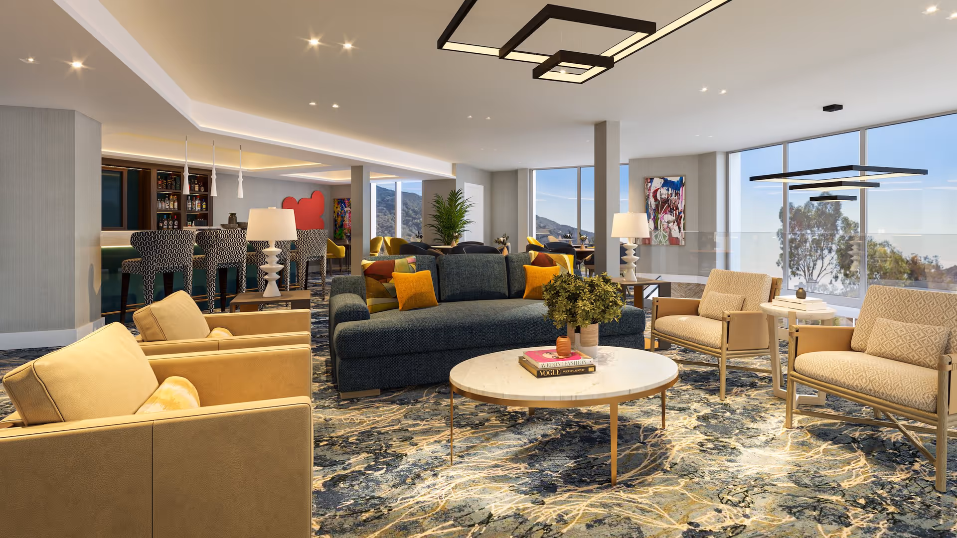 A spacious and modern senior living lounge area with comfortable seating including a dark blue sofa and beige armchairs arranged around a round white coffee table with books and a plant. Large windows provide natural light and a view of trees and hills outside. In the background, there is a bar area with patterned bar stools and pendant lighting. The room features contemporary lighting fixtures and colorful artwork on the walls.