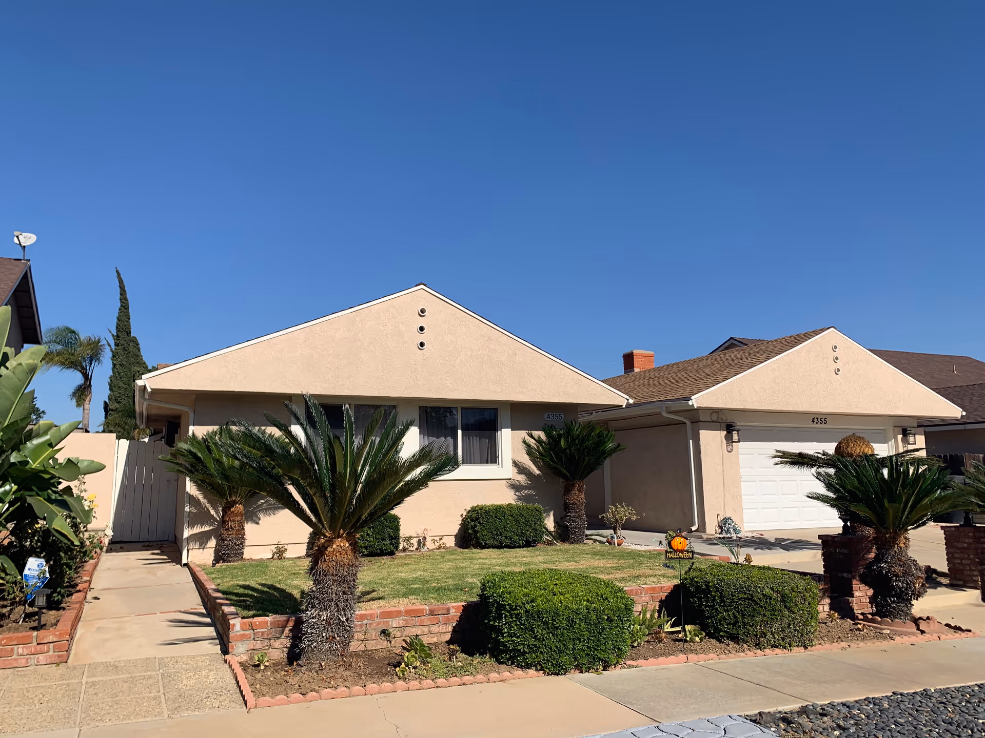 Front exterior view of a single-story beige house with a well-maintained lawn, small palm trees, and bushes. The house has a two-car garage and a clear blue sky in the background.