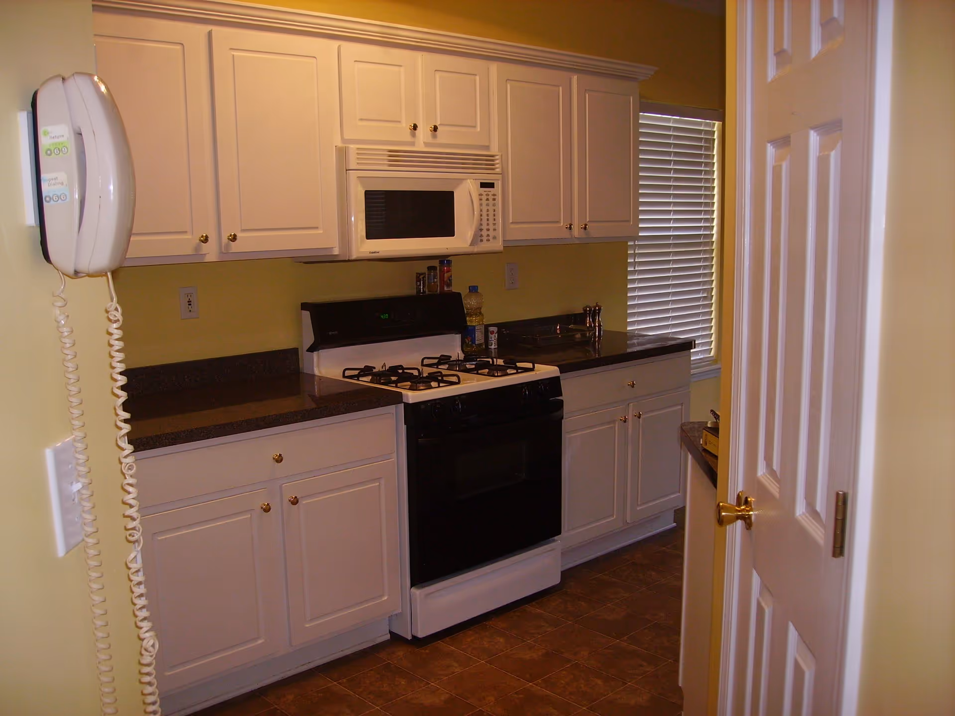 A small kitchen with white cabinets, a black and white gas stove, a white microwave above the stove, and a window with closed blinds. There is a wall-mounted white telephone on the left side and a partially open white door on the right.