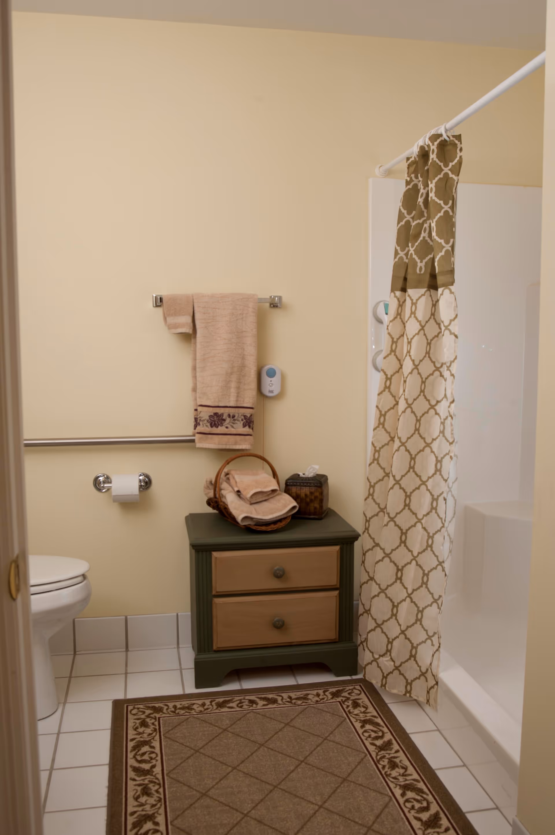 A bathroom with a white toilet, a towel rack holding a beige towel with a floral border, a small green cabinet with two drawers holding a basket of folded towels and a tissue box, a shower with a beige and white patterned curtain, and a beige and brown patterned rug on the tiled floor.