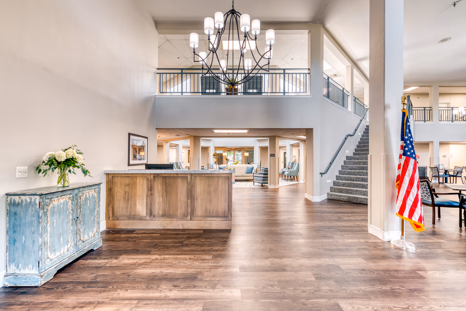 Spacious senior living facility interior with a wooden reception desk, a blue distressed cabinet with a vase of flowers, a large chandelier hanging from the ceiling, an American flag on a stand, carpeted stairs leading to an upper level, and seating areas with chairs and tables in the background.