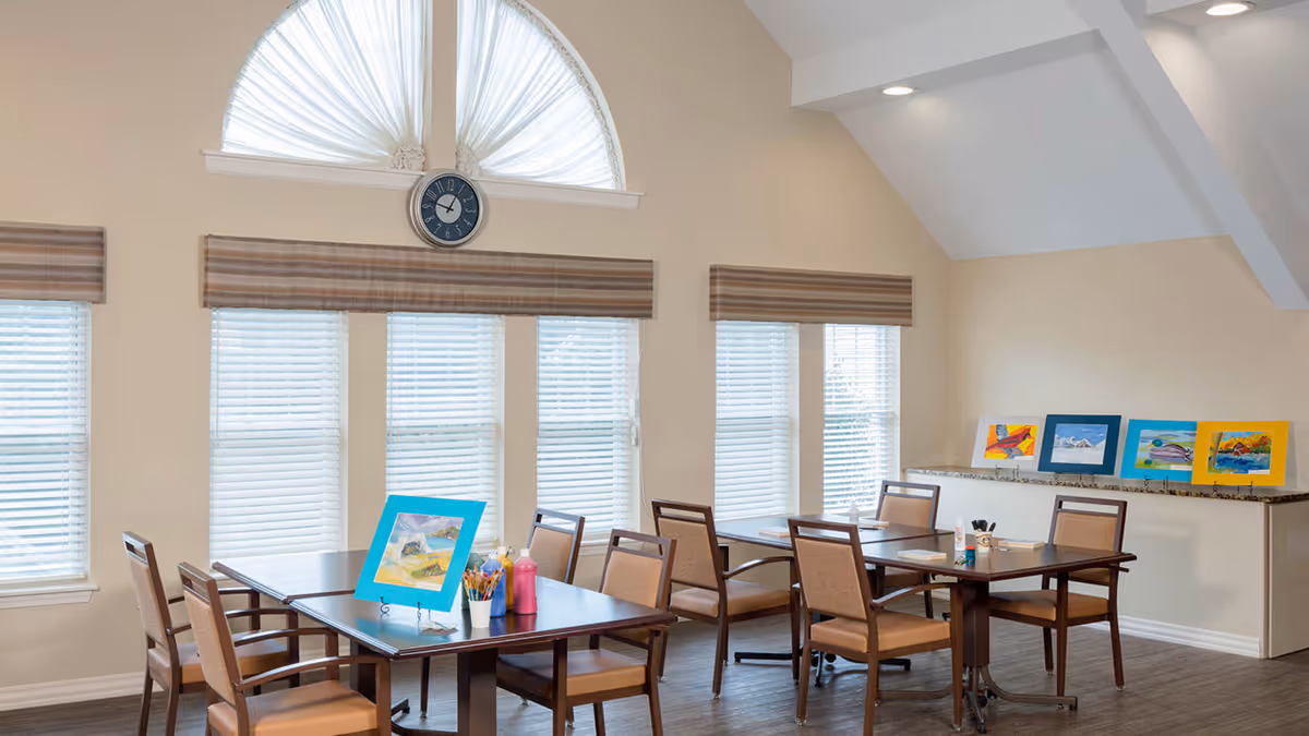 A bright room with multiple windows covered by blinds and a half-circle window at the top. There are two wooden tables with chairs around them. On the tables and countertop are colorful paintings and art supplies, suggesting an art or activity room.