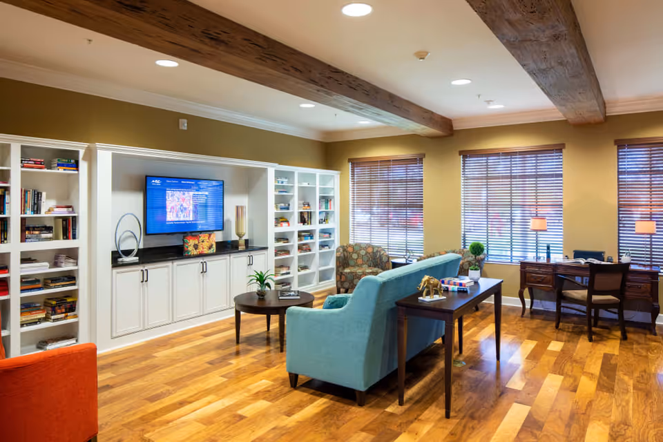 A cozy living room area in a senior living facility featuring a light blue sofa, a patterned armchair, and a wooden desk with a chair near three windows with blinds. The room has wooden floors, exposed wooden ceiling beams, built-in white bookshelves filled with books, and a flat-screen TV mounted on the wall.
