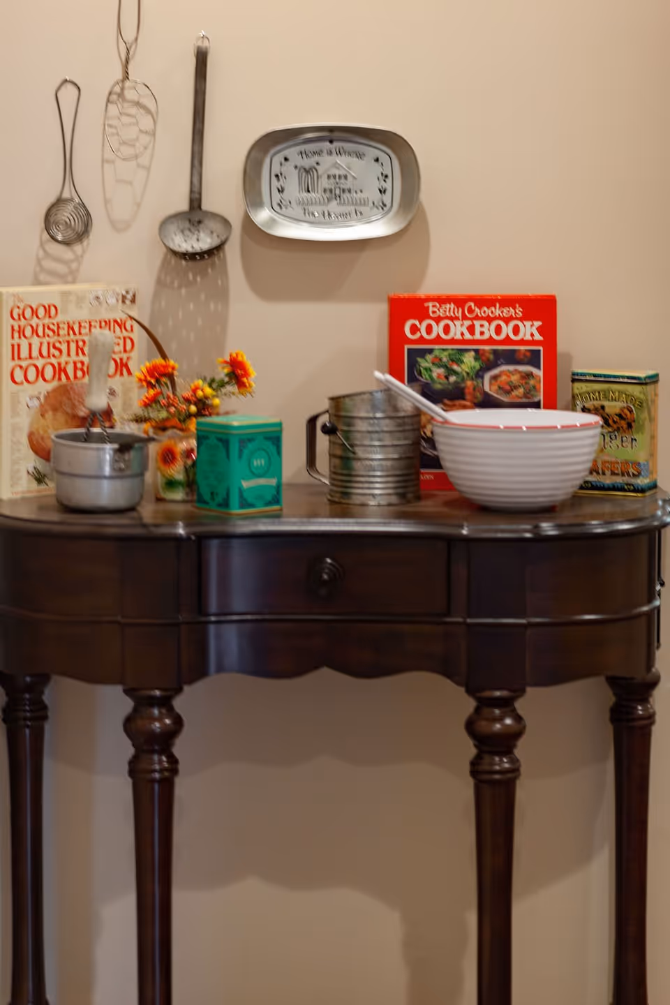 A wooden table with vintage kitchen items including two cookbooks, a metal sifter, a mixing bowl with a spoon, a small pot, a green tin, and a small vase with orange flowers. Above the table, three metal kitchen utensils hang on the wall along with a decorative metal tray with the phrase 'Home is Where The Heart Is'.