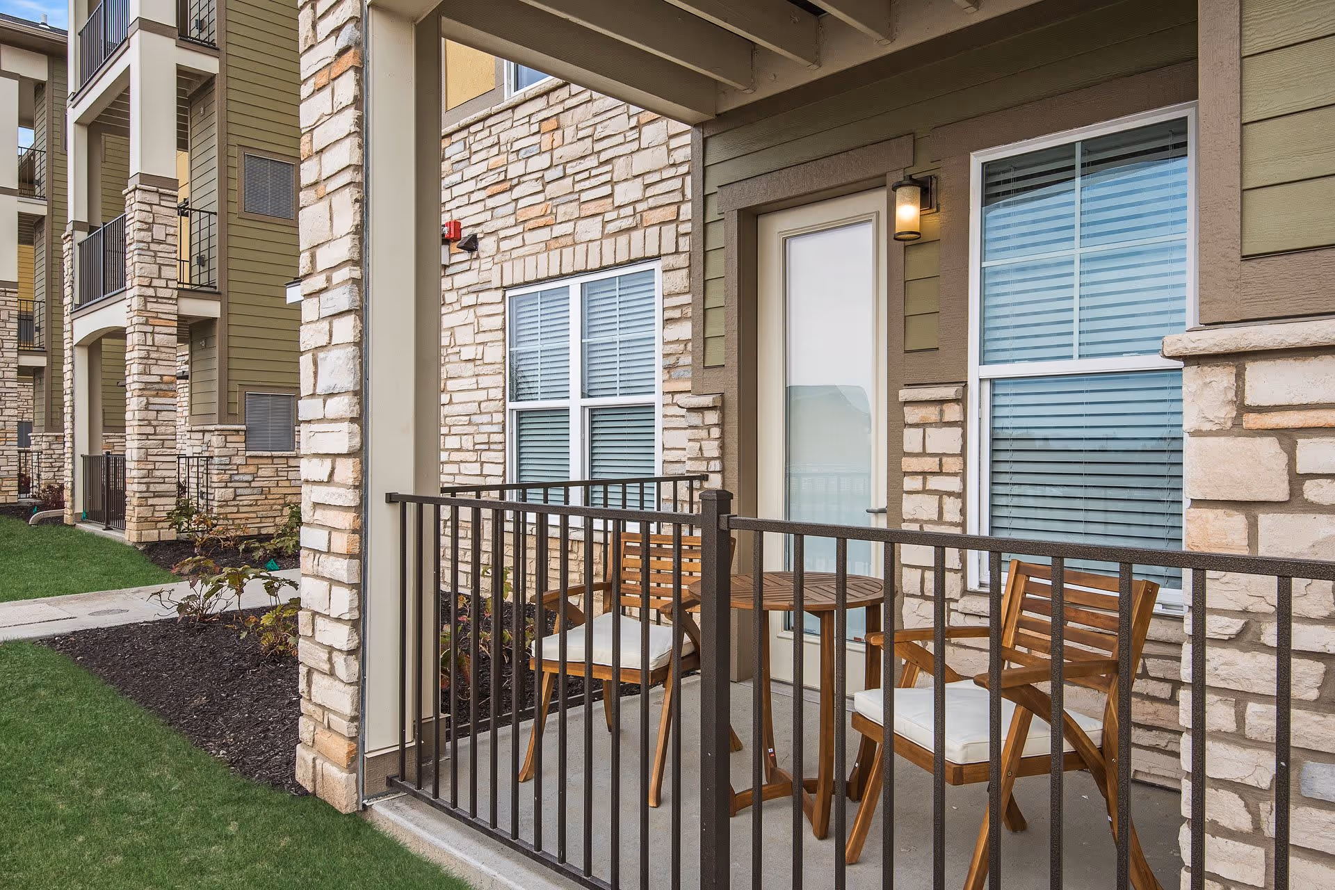 A small outdoor patio area with two wooden chairs with white cushions and a round wooden table. The patio is enclosed by a black metal railing and is attached to a building with stone and green siding exterior walls. There are two windows with white blinds and a door with frosted glass leading inside. The surrounding area has green grass and some landscaping with mulch and plants.