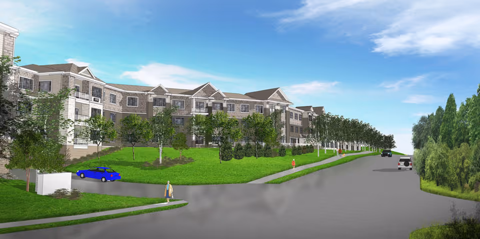 Rendering of a large senior living facility building with multiple floors, surrounded by green lawns and trees. A few cars are parked on the road in front of the building, and a few people are walking on the sidewalks. The sky is clear with some clouds.