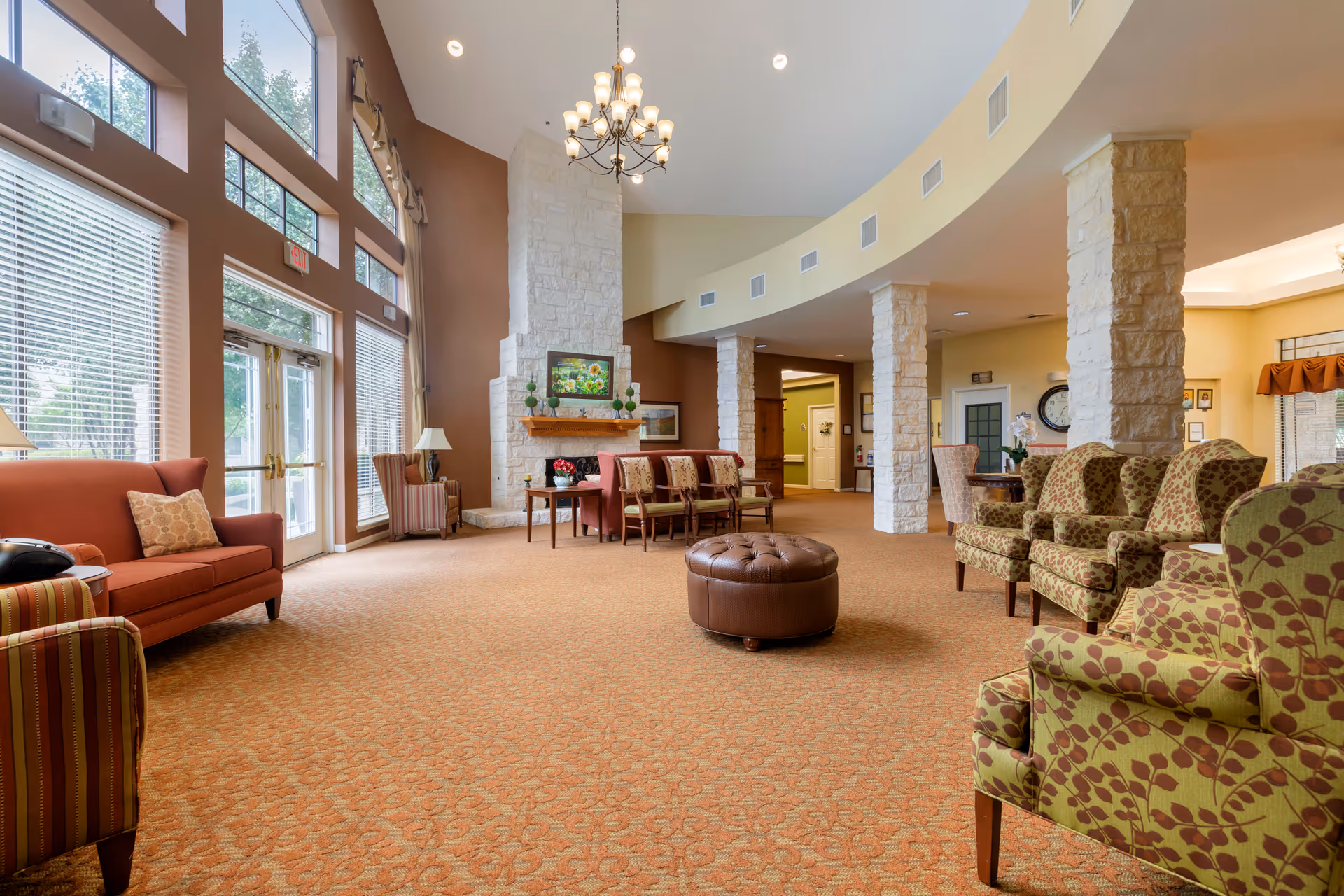 Spacious senior living facility common area with high ceilings, large windows, and a stone fireplace. The room is furnished with various upholstered chairs, sofas, and a round leather ottoman. Warm lighting and earth-toned decor create a welcoming atmosphere.