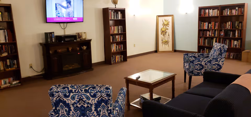A cozy living room area with two blue patterned armchairs, a dark sofa, a glass-top wooden coffee table, and bookshelves filled with books. A flat-screen TV is mounted on the wall above a dark wooden electric fireplace. There is a framed floral artwork leaning against the wall in the corner.