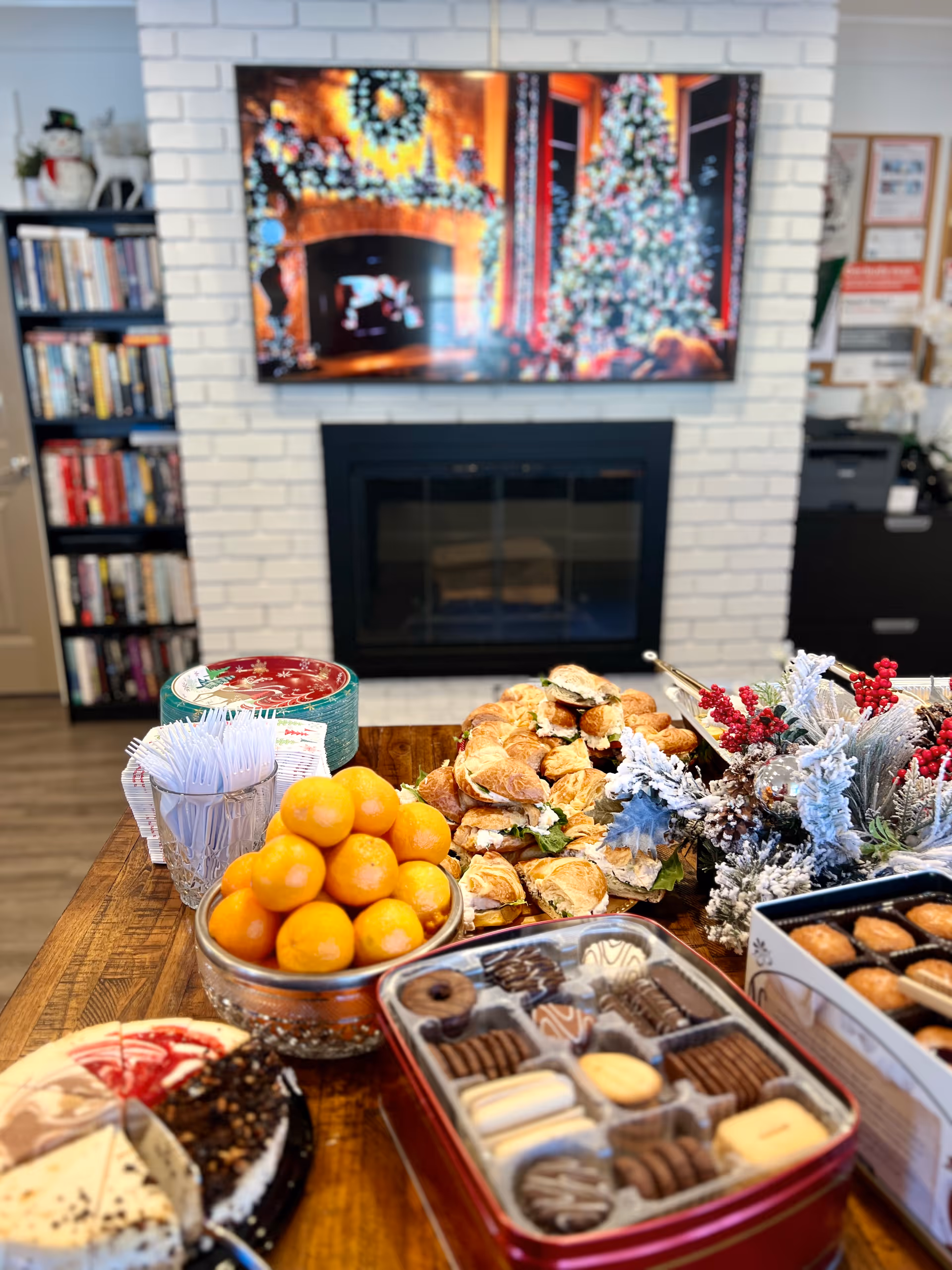 A wooden table set with a variety of food including a bowl of oranges, croissant sandwiches, assorted cookies in a tin, and a partially sliced cake. In the background, there is a white brick fireplace with a TV above it displaying a festive holiday scene with a Christmas tree and wreath. To the left, a bookshelf filled with books and a snowman decoration is visible. To the right, there are some holiday floral decorations and office supplies.