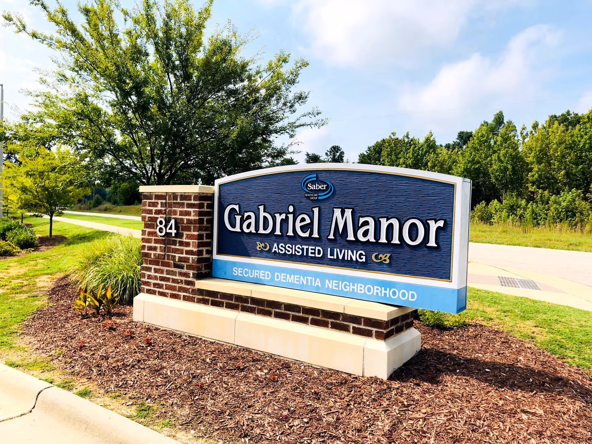 Entrance sign for Gabriel Manor Assisted Living reading 'Secured Dementia Neighborhood' on a landscaped roadside.
