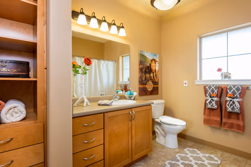 A clean and well-lit bathroom with wooden cabinets, a countertop with a sink, a mirror above the sink, a toilet, and a window with two decorative towels hanging below it. There is a white rolled towel on a wooden shelf, a vase with a red rose on the countertop, and a painting on the wall next to the toilet. The floor has a patterned rug.