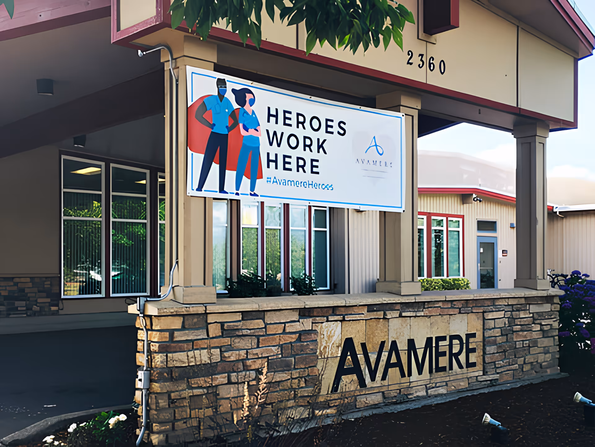 Exterior entrance of Avamere Rehabilitation of Eugene with a stone sign displaying 'AVAMERE' and a banner above that reads 'HEROES WORK HERE' featuring illustrated healthcare workers.