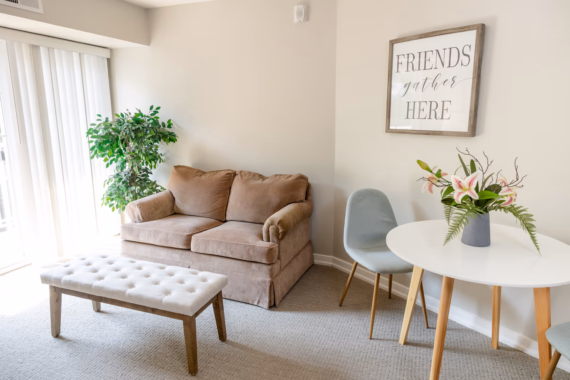 A cozy living room area with a beige loveseat, a tufted bench, a small round white table with wooden legs, a gray chair, a vase with flowers on the table, a potted green plant near a window with vertical blinds, and a framed wall art that reads 'FRIENDS gather HERE'.