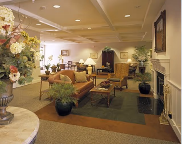 Warm, carpeted common lounge with sofas, armchairs, coffee tables, potted plants, lamps, and a fireplace.