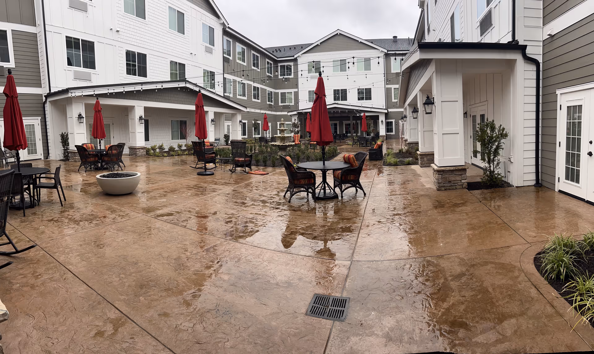 Wet courtyard patio of a multi-story assisted living community with tables, chairs and red umbrellas.