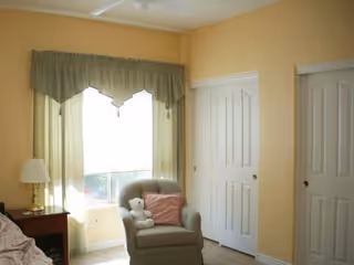 A cozy bedroom corner with a beige armchair holding a white teddy bear and a pink pillow, next to a window with light green curtains. There is a wooden nightstand with a lamp beside the chair, and two white closet doors on the adjacent wall.