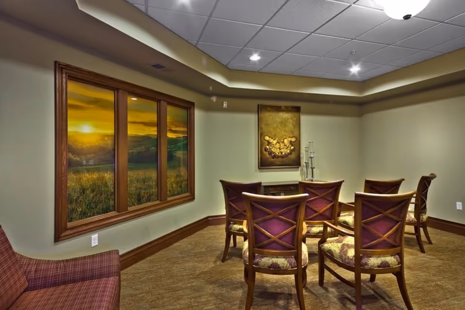 A small meeting or gathering room with six wooden chairs arranged in two rows facing a wall with a framed artwork and decorative glass candle holders. There is a large window on the left wall showing a scenic sunset over a field and hills. The room has beige walls, a patterned carpet, and a drop ceiling with recessed lighting.