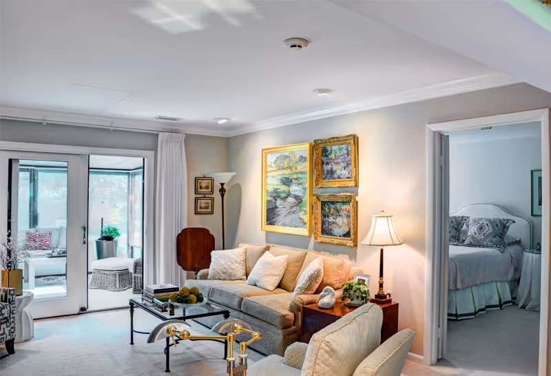 Bright living room with a beige sofa, glass coffee table, wall paintings, lamp, and open doorways to a bedroom and a sunroom.