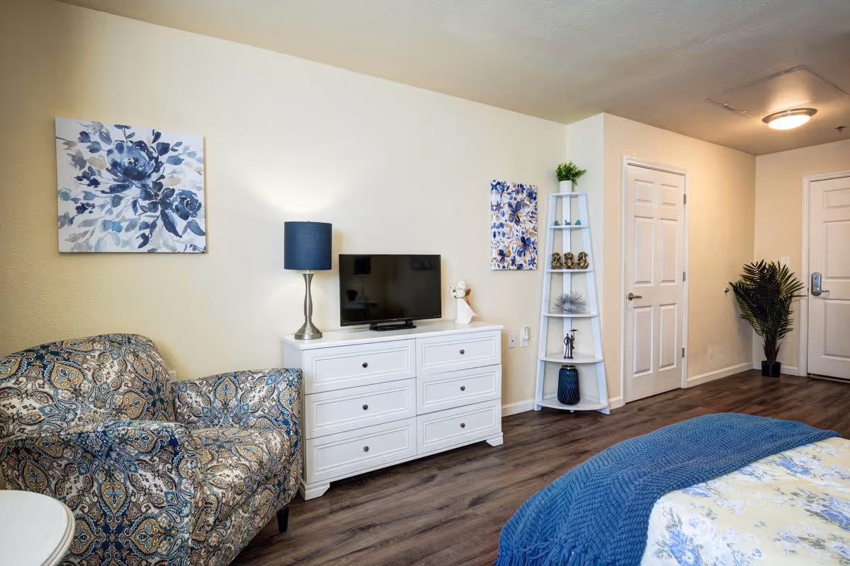 A cozy bedroom in a senior living facility featuring a patterned armchair, a white dresser with a TV and lamp on top, floral artwork on the walls, a corner shelf with decorative items, a bed with a blue blanket, and a potted plant near the door.
