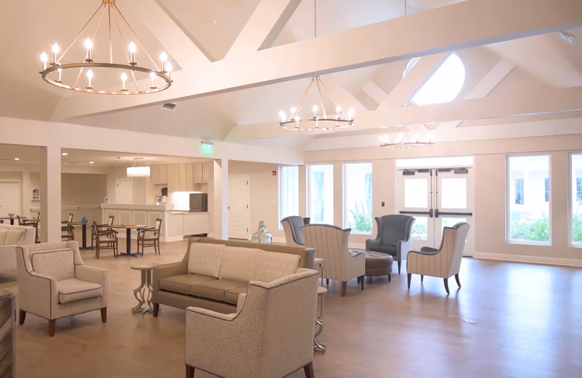 A spacious and well-lit common area in a senior living facility featuring multiple seating arrangements including sofas, armchairs, and small tables. The room has high vaulted ceilings with large circular chandeliers, several windows allowing natural light, and a double door exit. In the background, there is a kitchenette area with cabinets and a microwave.