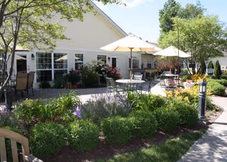Outdoor patio area with tables, chairs, and umbrellas surrounded by well-maintained garden beds with various plants and flowers. The patio is adjacent to a light-colored building with multiple windows, and there are trees providing shade.