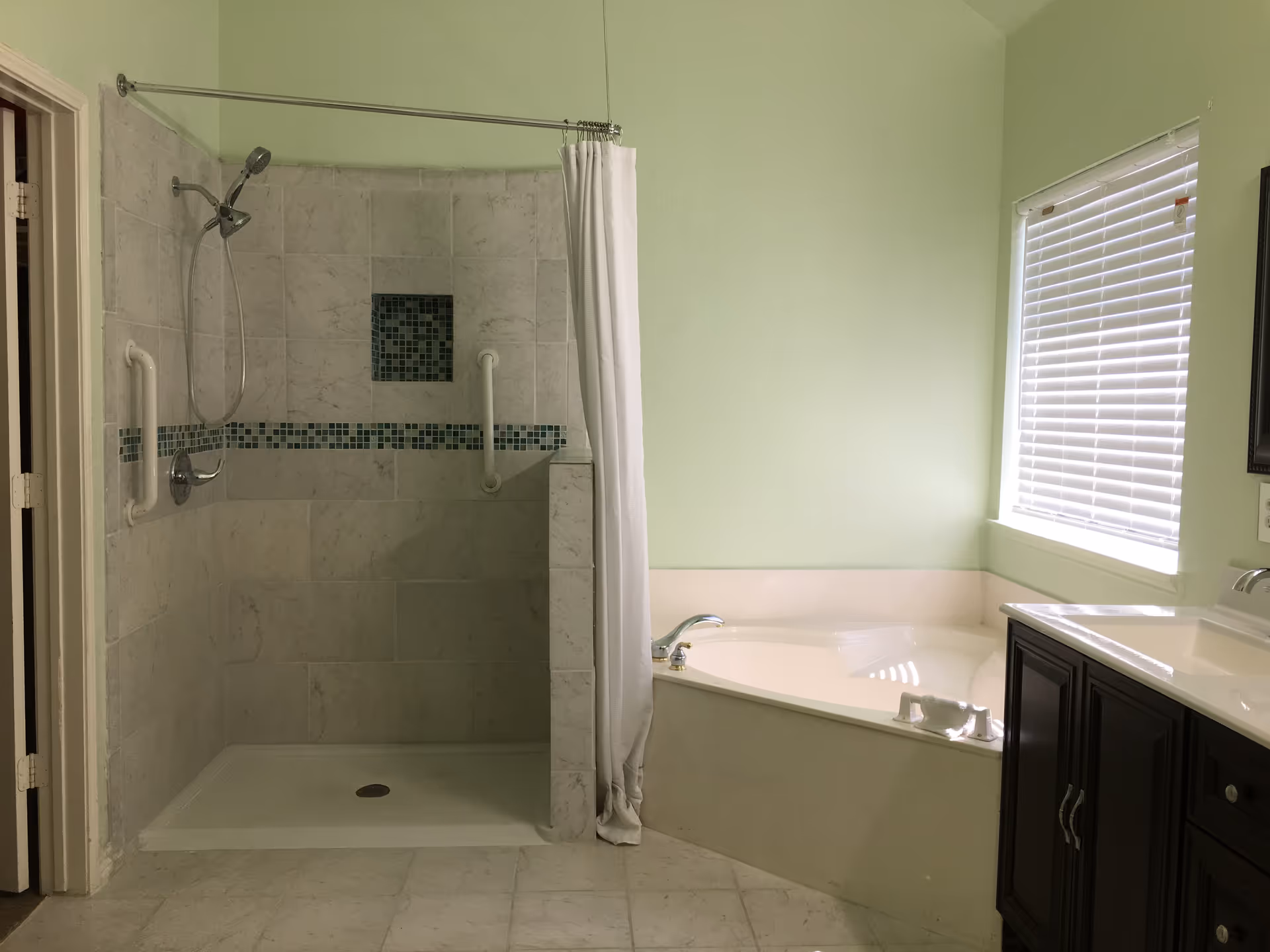 A bathroom with a walk-in shower featuring white and gray tiles with blue mosaic accents, grab bars, and a white shower curtain. Next to the shower is a corner bathtub with a faucet, and a window with white blinds above it. There is also a dark wood vanity with a white sink on the right side.
