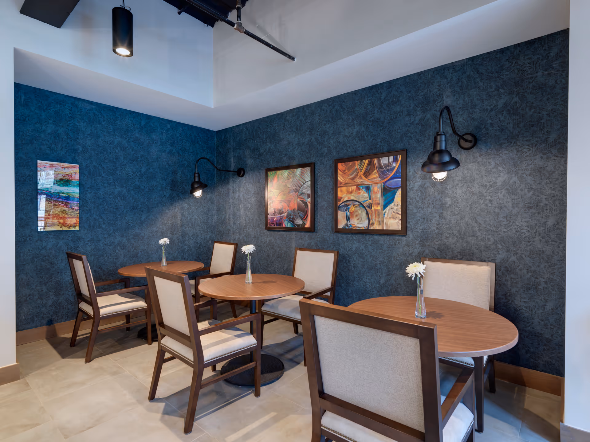 Small dining area with three round wooden tables, upholstered chairs, single-flower vases, a textured dark blue wall with framed artwork and industrial wall lamps.
