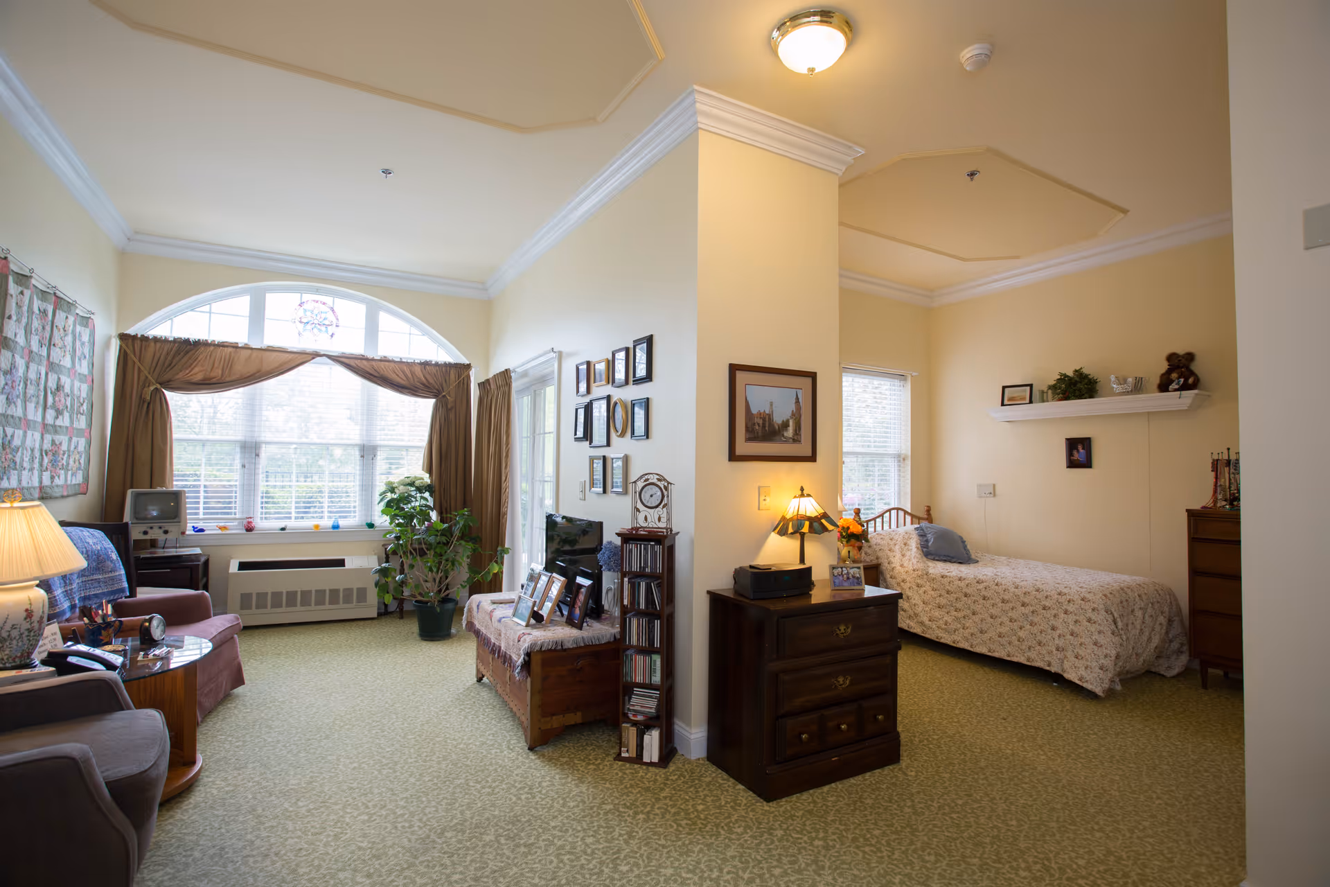 A cozy senior living room and bedroom area separated by a partial wall. The living room features comfortable chairs, a small TV, a large window with curtains, and various decorations including plants and framed pictures. The bedroom area has a single bed with a floral bedspread, a nightstand with a lamp, a dresser, and shelves with decorative items.