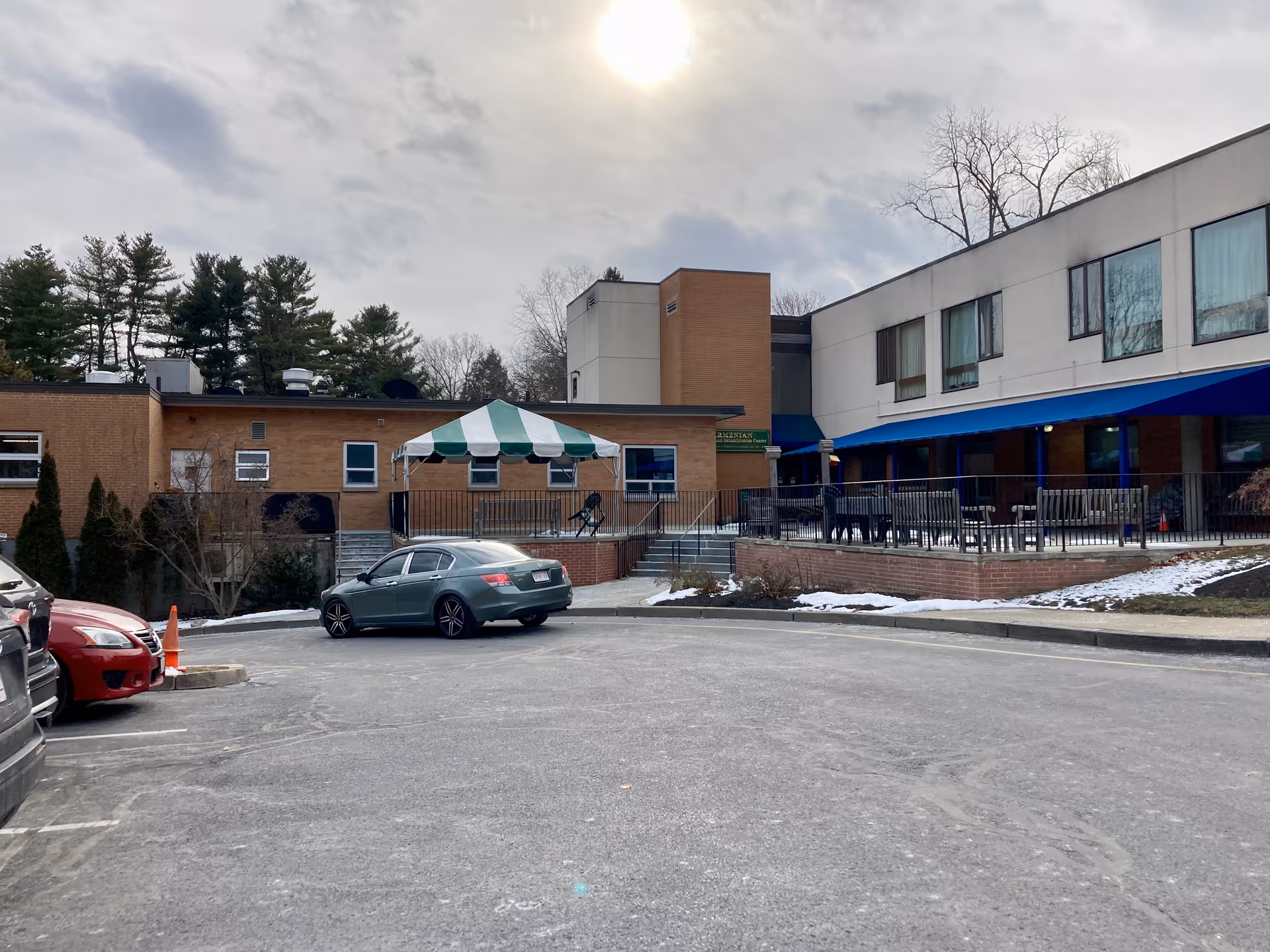 Exterior view of Armenian Nursing & Rehabilitation Center showing a parking lot with several cars, a building with brick and white walls, windows with curtains, a blue awning, and a green and white striped canopy over a small patio area. Some snow is visible on the ground and trees are in the background under a cloudy sky.