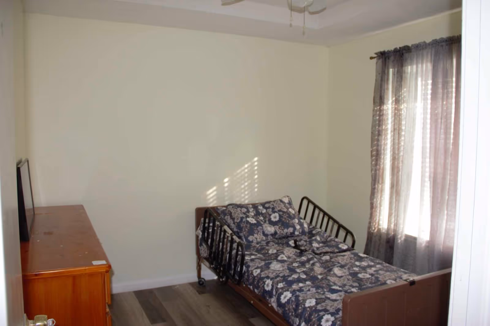 Small bedroom with a single hospital-style bed with floral bedding, a wooden dresser, and a curtained window.