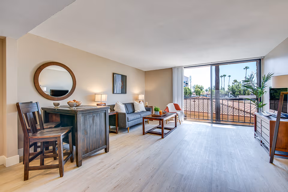 A bright living room with light wood flooring, featuring a dark wooden dining table with two chairs on the left, a gray sofa with white pillows and two side tables with lamps, a wooden coffee table, and a TV on a wooden stand to the right. Large sliding glass doors open to a balcony with a view of palm trees and rooftops outside.