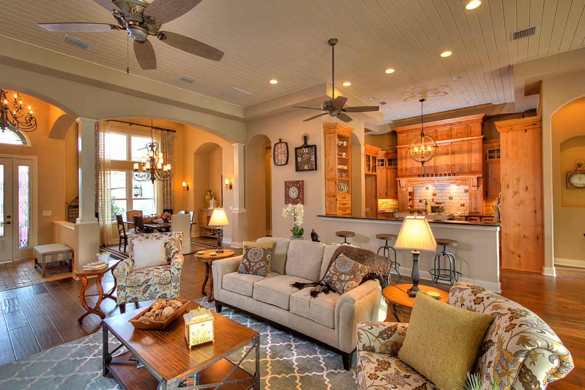 A spacious and warmly lit living room with beige and floral patterned armchairs, a beige sofa with decorative pillows, a wooden coffee table with a decorative tray and lantern, and two table lamps. The room features ceiling fans, wooden flooring, and an open layout leading to a kitchen with wooden cabinetry and a breakfast bar with stools. In the background, a dining area with a chandelier and large windows is visible.