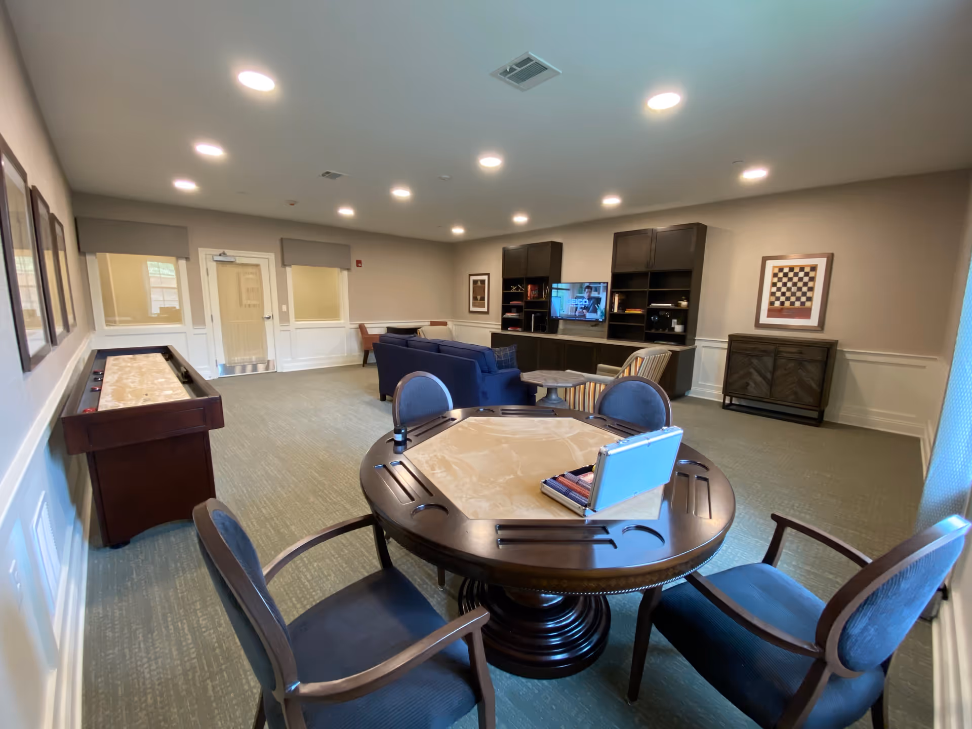 A spacious senior living common area with a round game table and four chairs in the foreground, a shuffleboard table along the left wall, and a seating area with a blue sofa, armchairs, and a TV mounted on a dark wood entertainment center in the background. The room has beige walls, carpeted floor, recessed ceiling lights, and framed artwork on the walls.