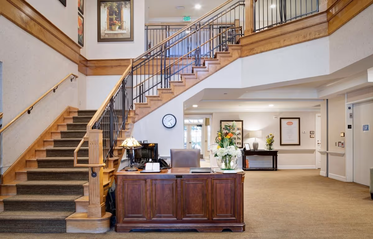 Lobby reception area with a wooden desk, floral arrangement, and a grand staircase leading to the second floor.